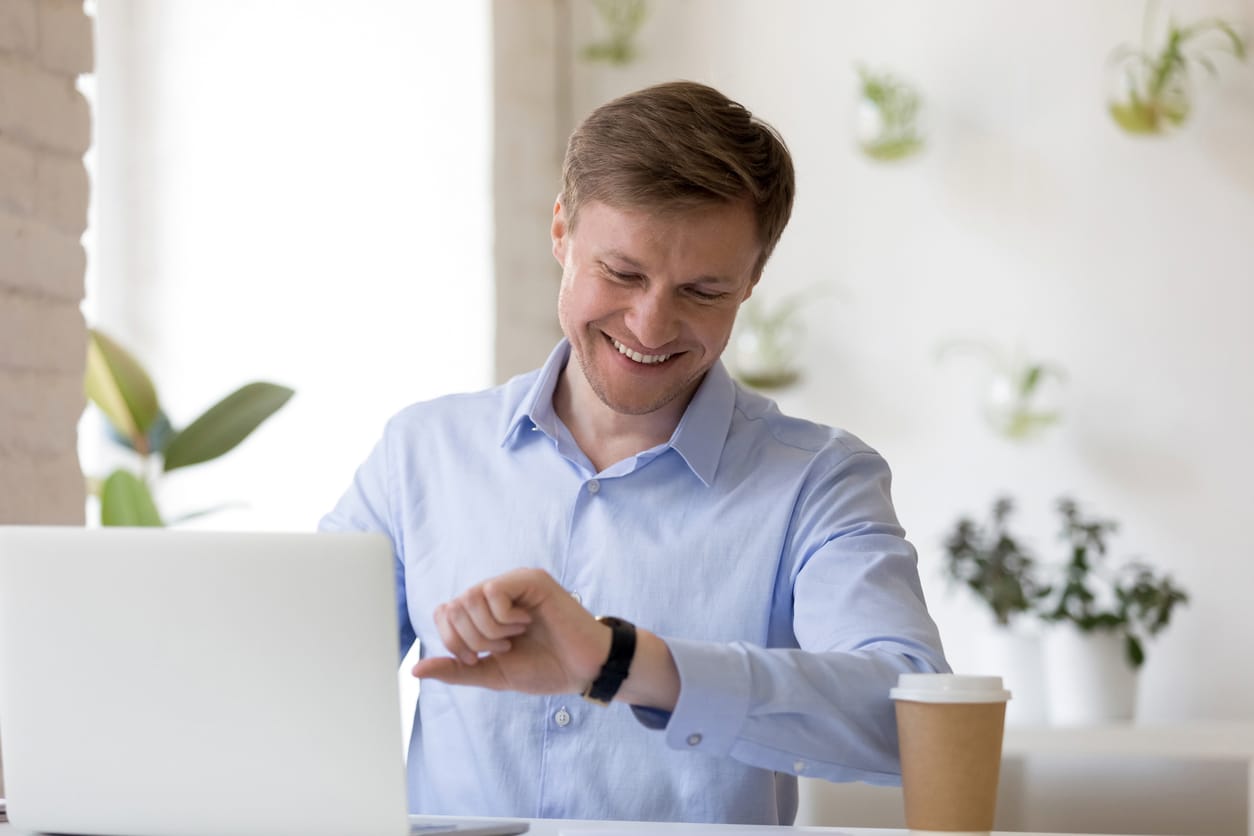 Arbeitnehmer vor Laptop blickt lächelnd auf die Uhr